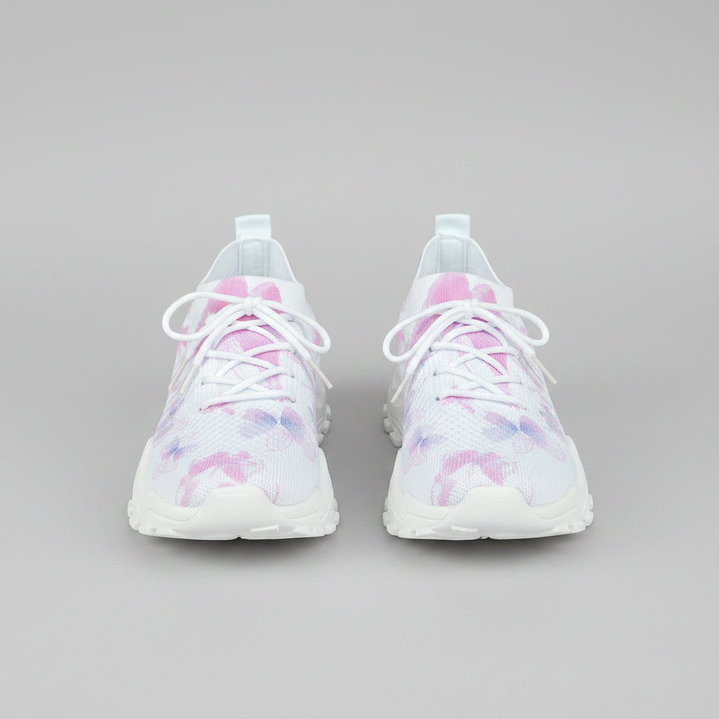 White sneakers with pink floral patterns worn by a person sitting on a woven chair.